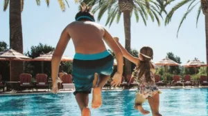 Dos niños saltando a una piscina rodeada de palmeras en un día de sol